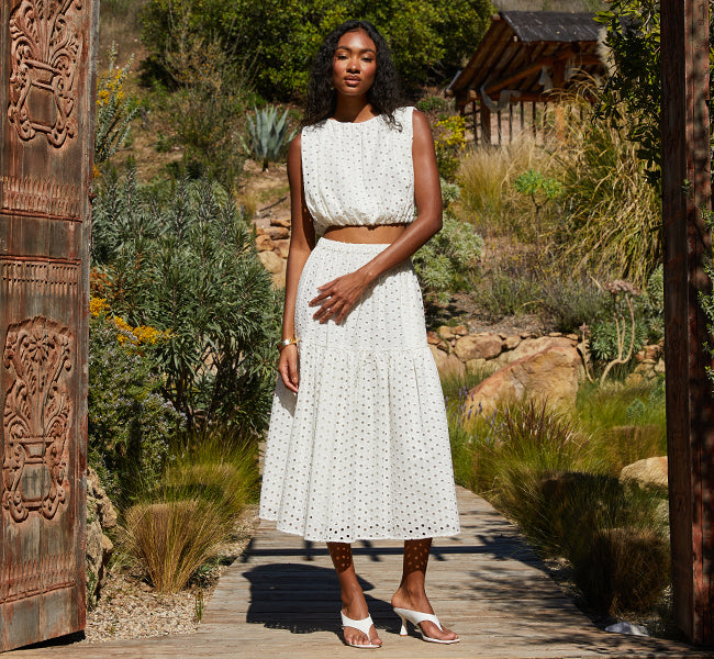 Model wearing a white eyelet crop top and matching midi skirt, standing on a wooden path surrounded by desert plants and rustic architecture.
