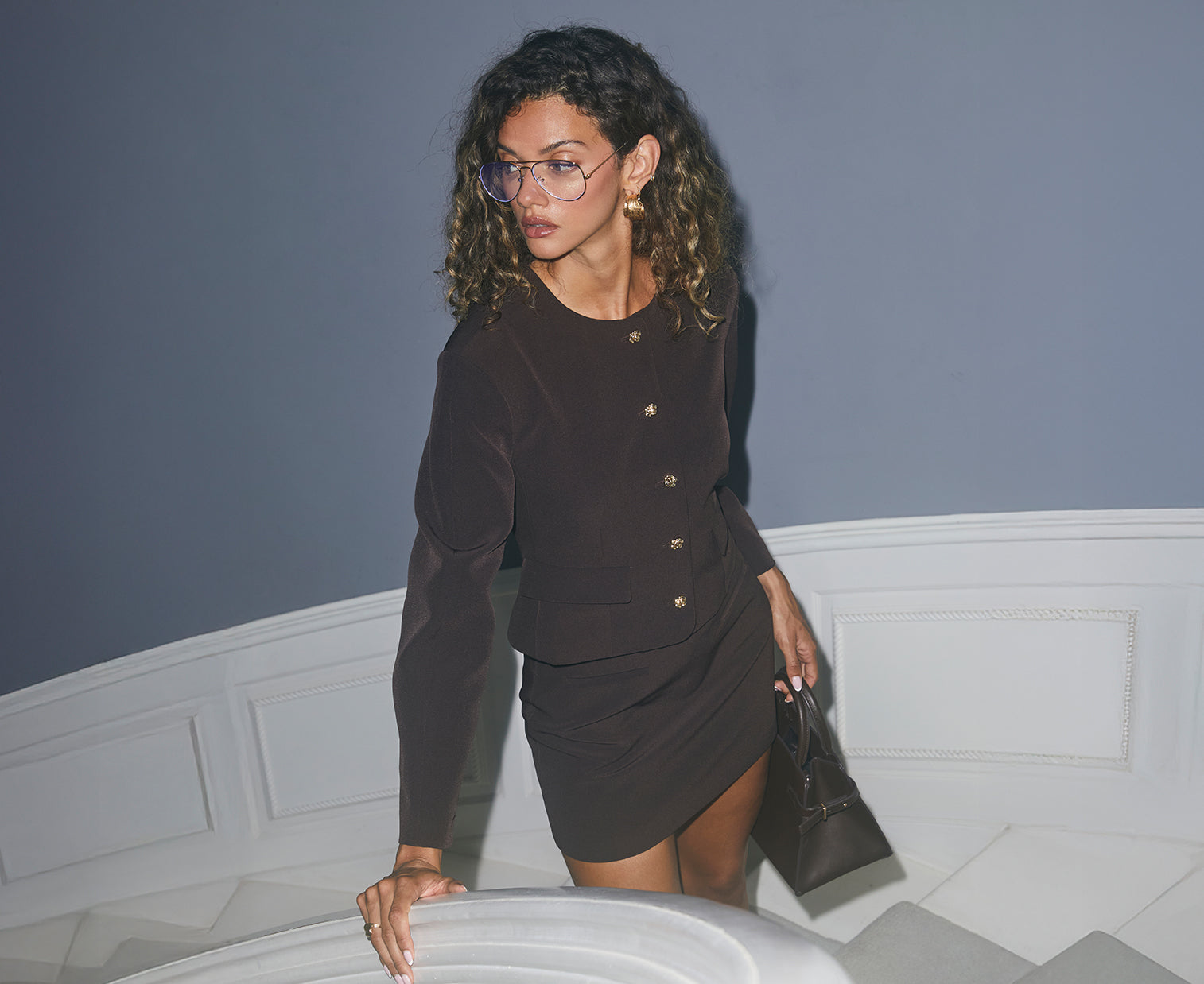 A woman is wearing a black, fitted blazer with gold buttons down the front, paired with a matching black skirt that is slightly asymmetrical. She accessorizes with large gold earrings, glasses, and holds a small black handbag.