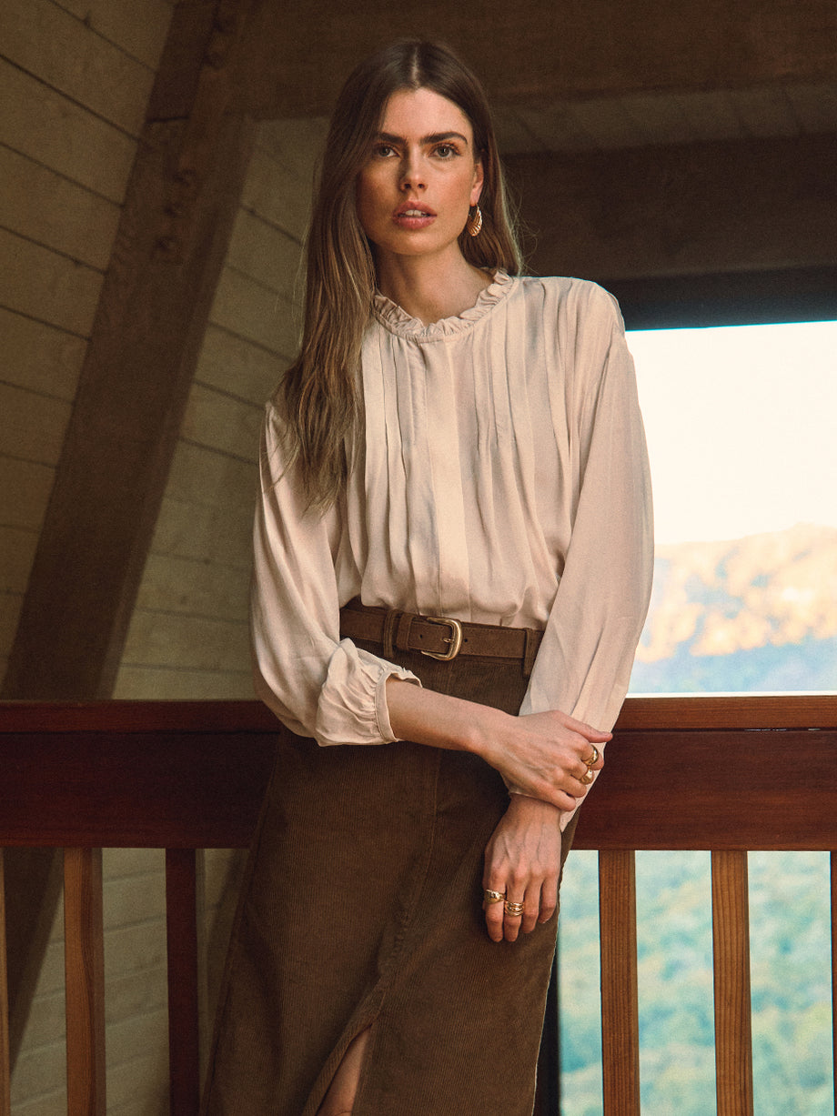 Woman in a beige blouse and brown skirt standing on a balcony with a scenic view.