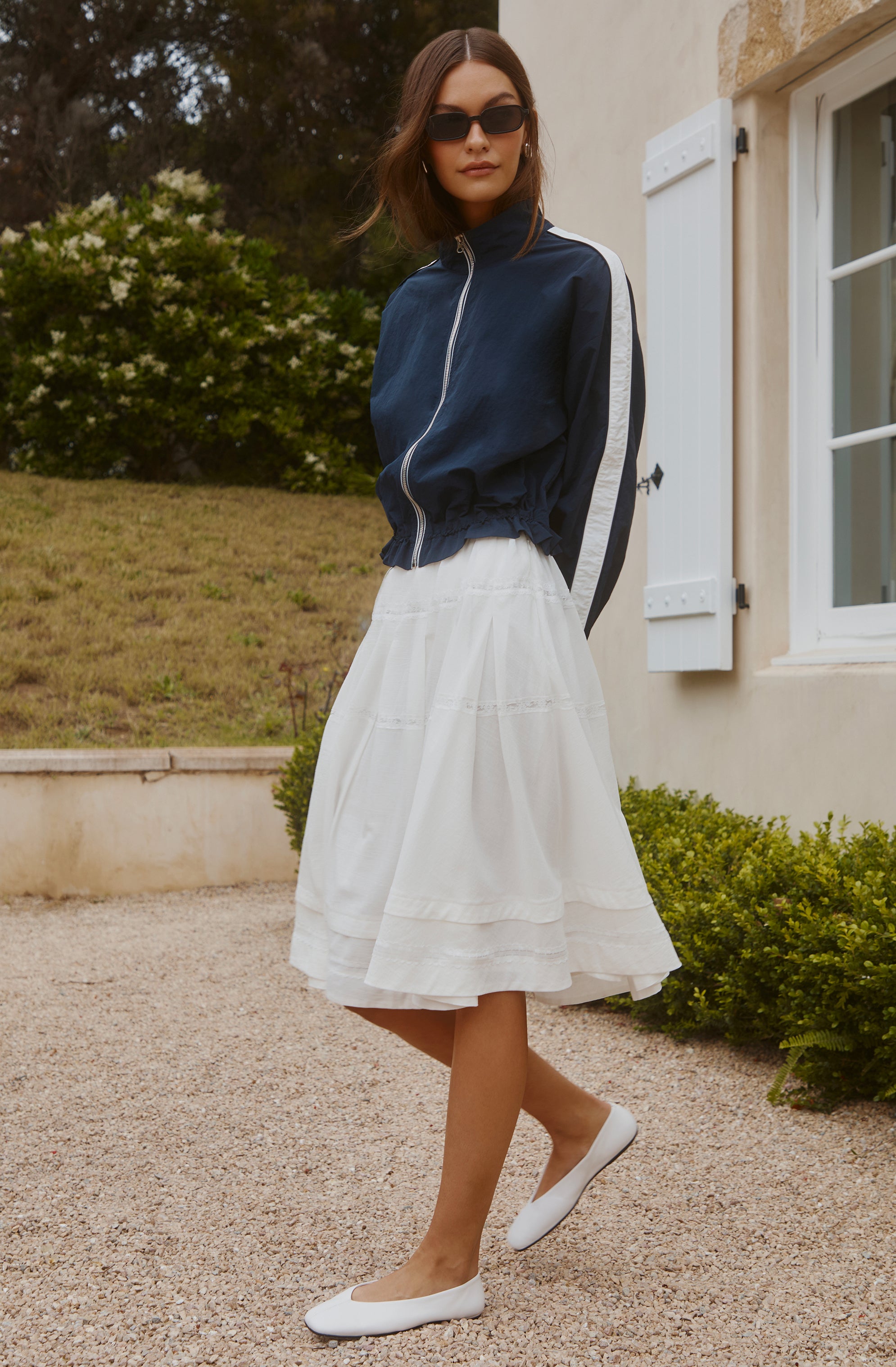 Woman wearing a navy blue zip-up jacket with white and black stripes, paired with a white midi skirt and white flats.