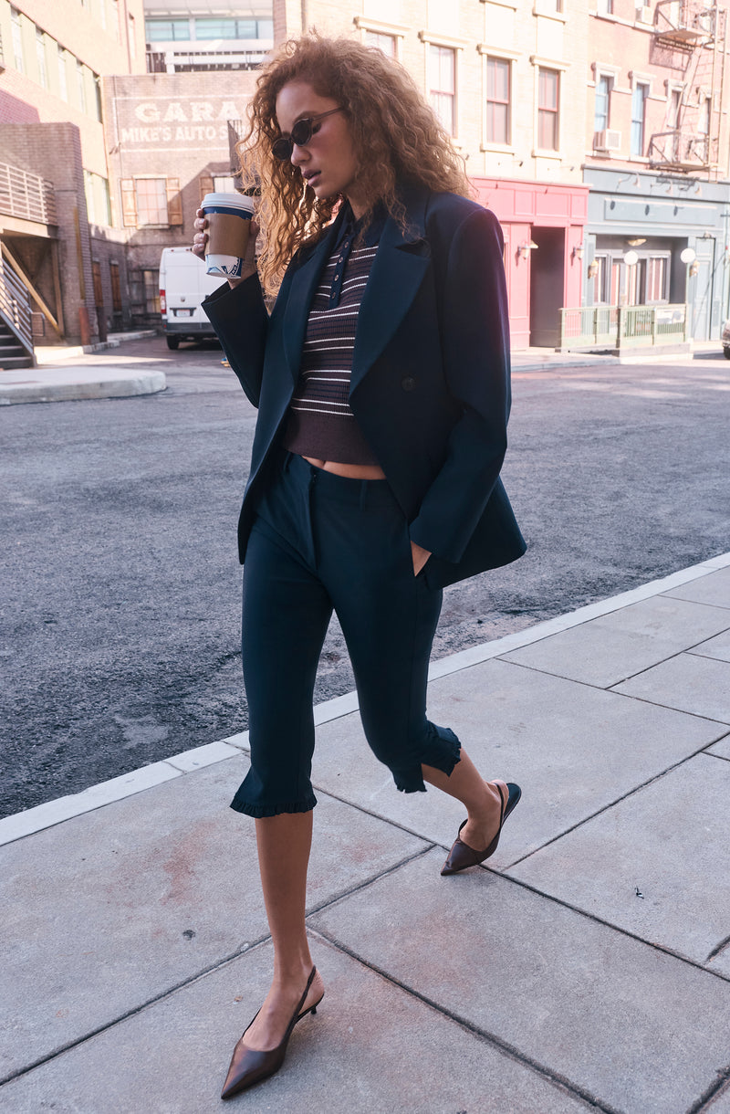 Woman wearing a black blazer, striped crop top, cropped dark jeans with ruffled hems, and pointed heels while holding a coffee cup.