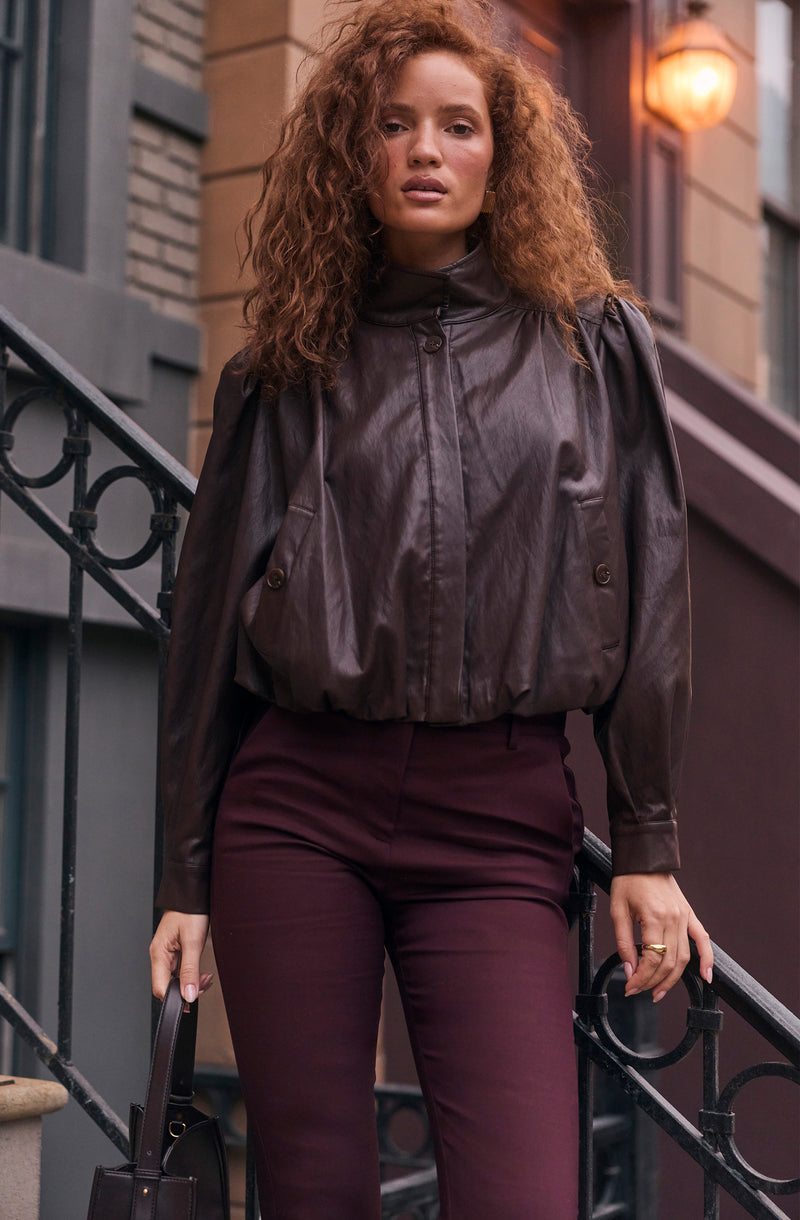 Woman in a dark leather jacket and burgundy pants, holding a black handbag, standing outside.