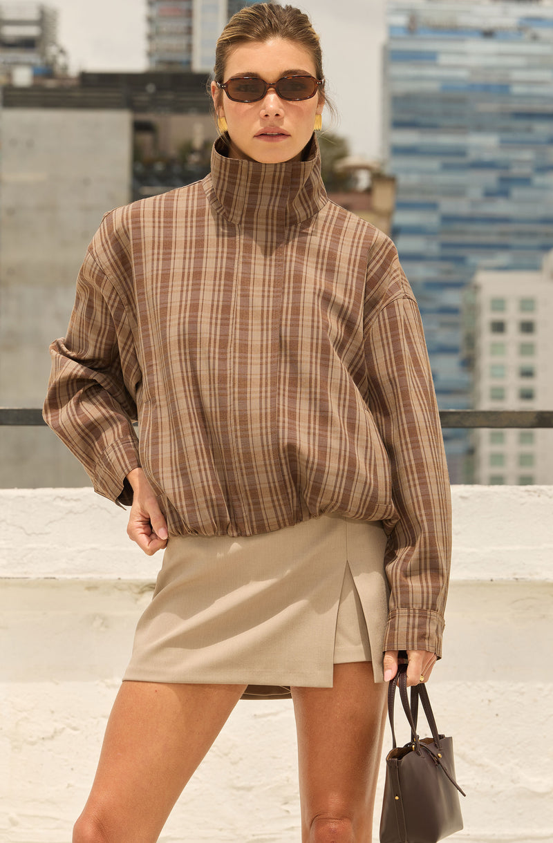 Woman wearing a plaid brown high-neck jacket, beige mini skirt, and sunglasses on a rooftop.