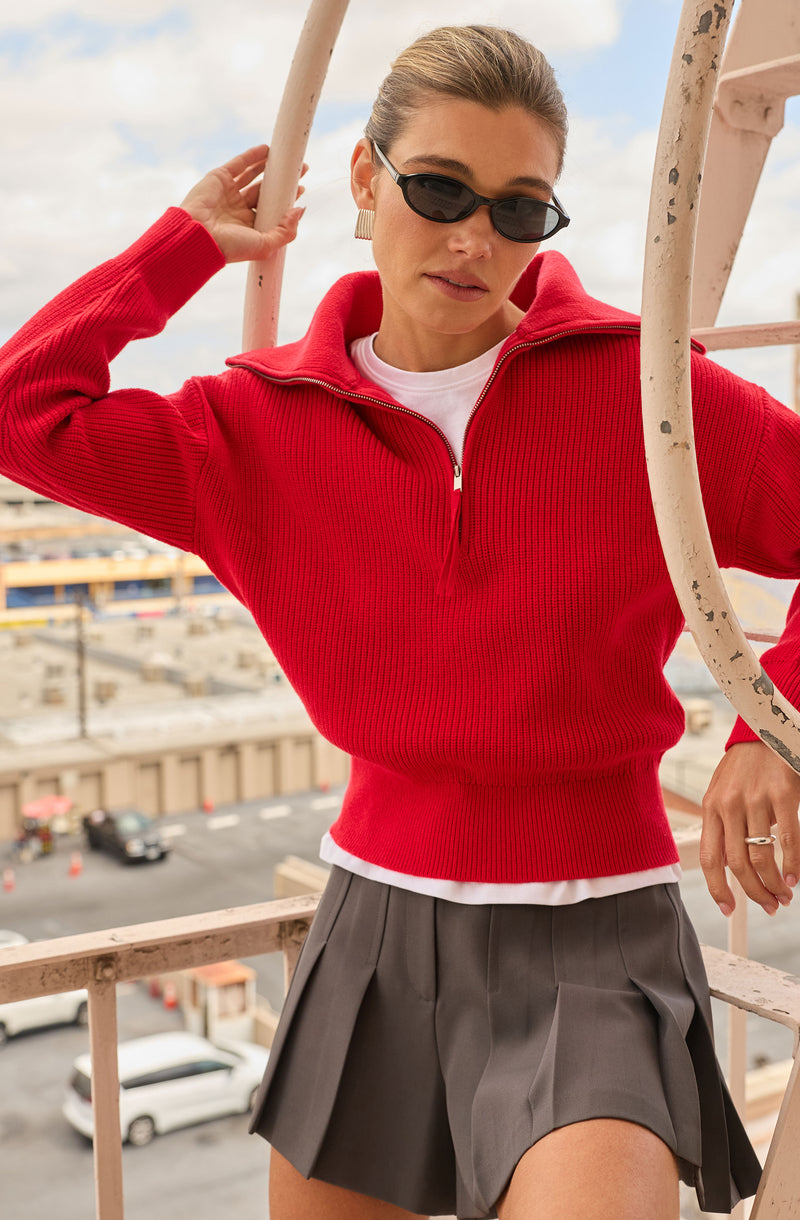 Woman wearing a red ribbed zip-up sweater, white shirt underneath, and a short gray pleated skirt.