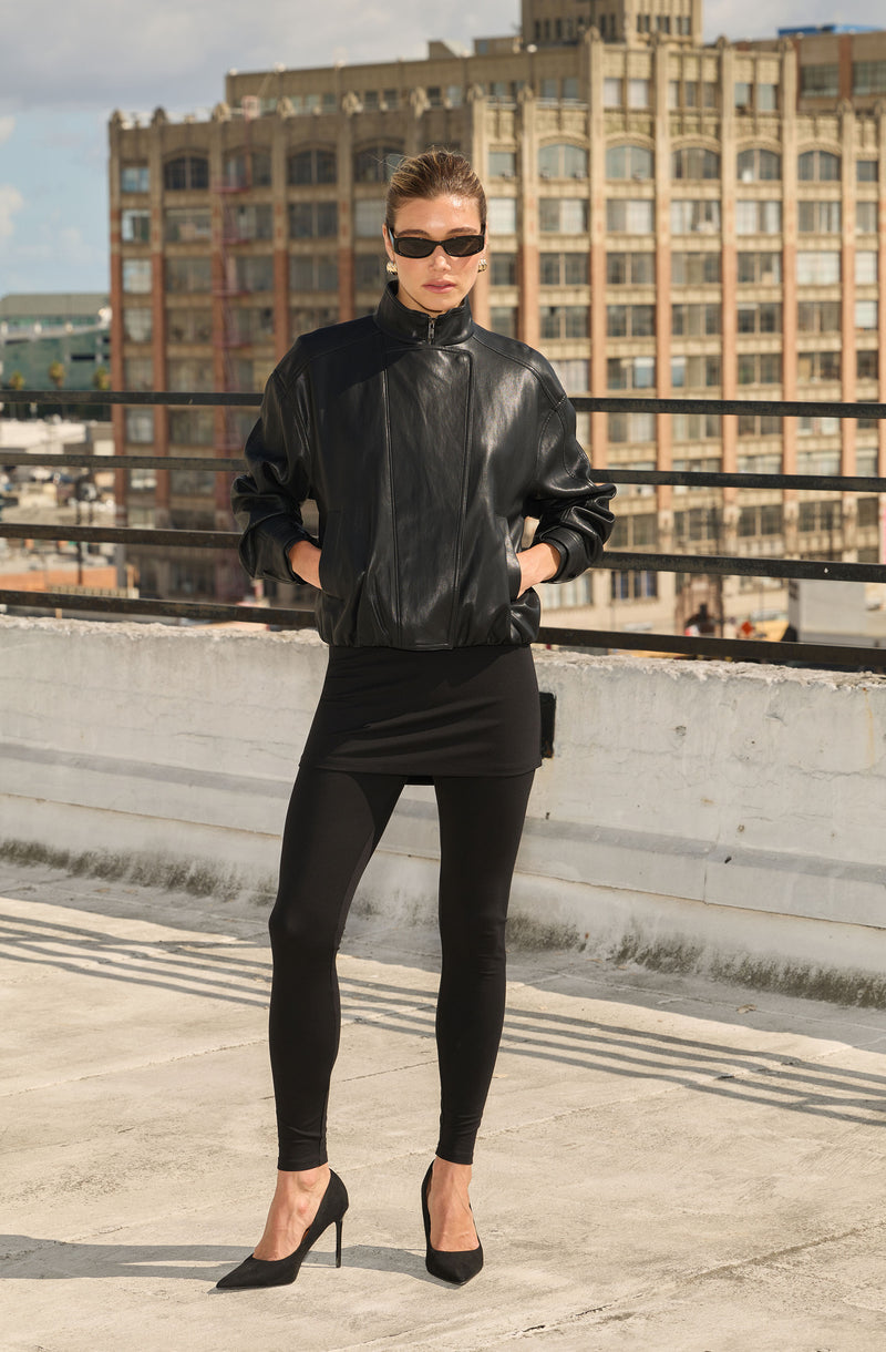 Woman in black leather jacket, leggings, high heels, and sunglasses on rooftop.