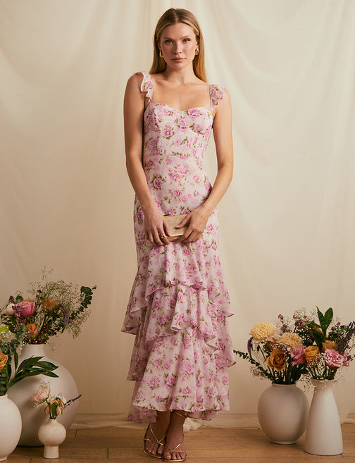 Woman in a pink floral dress with ruffles, holding a beige clutch, surrounded by flower vases.