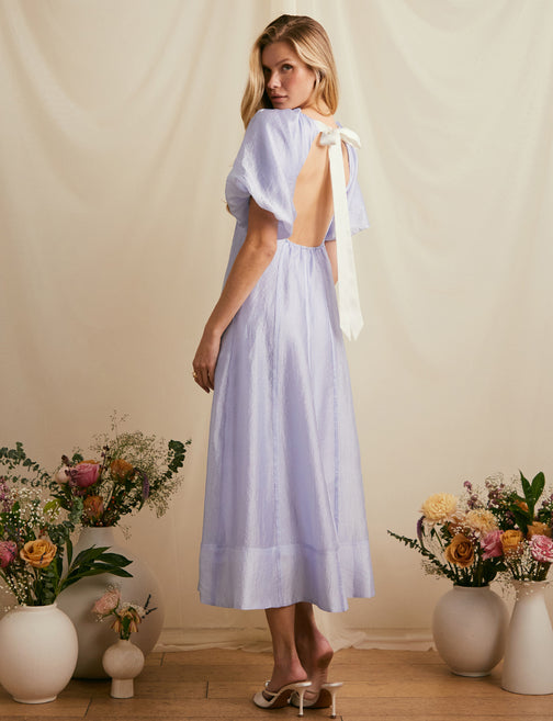 Woman wearing a light purple dress with puffed sleeves, open back with shoulder ties, and a loose, flowing skirt.