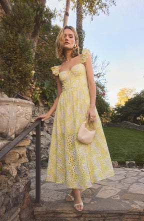 A woman is wearing a light yellow, floral-patterned maxi dress with ruffled cap sleeves and a fitted bodice. She is holding a small, light-colored bag with a gathered handle. She is also wearing open-toed, light-colored sandals.