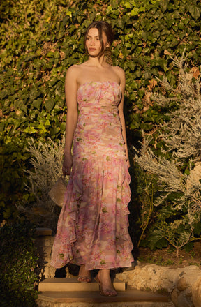 A woman is wearing a strapless, floor-length dress with a pastel floral pattern in shades of pink, peach, and green. The dress features a fitted bodice and a flowing skirt with ruffled detailing toward the bottom. She is holding a glittery clutch bag in her left hand.