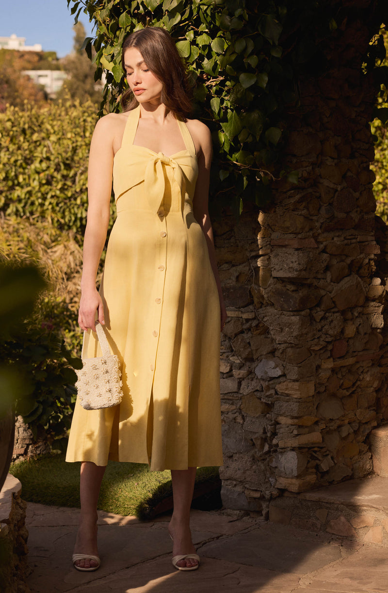 A woman is wearing a sleeveless, button-down, yellow midi dress with a large bow tie at the neckline. She is holding a small, white, beaded handbag and wearing open-toed beige sandals. The dress has a fitted bodice and flows loosely to her mid-calf.
