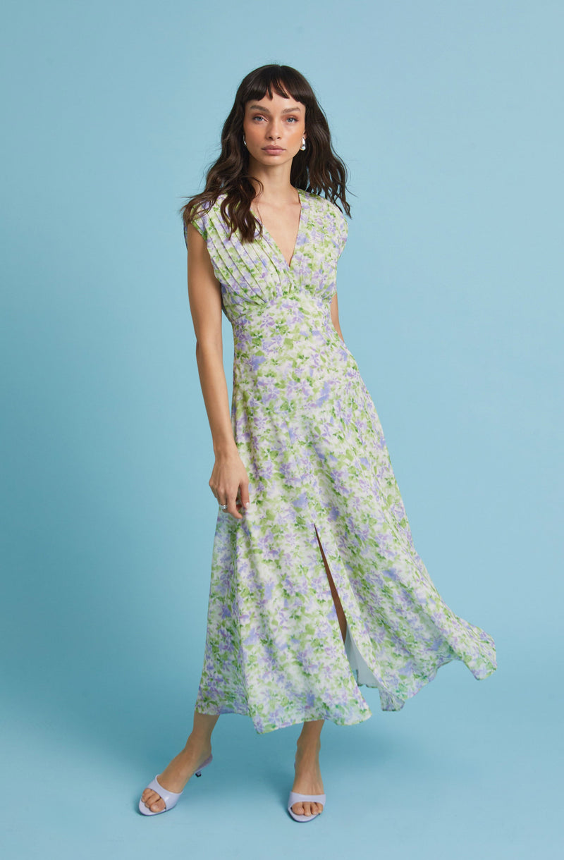 A woman is wearing a sleeveless, floor-length dress with a V-neckline. The dress features a floral pattern in shades of light purple, green, and white. It has a fitted waist and a front slit that reveals her legs. She is also wearing white open-toe heels.