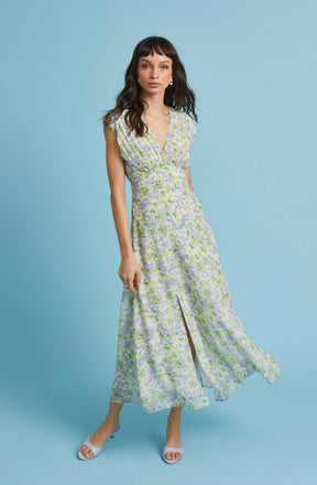 A woman is wearing a sleeveless, floor-length dress with a V-neckline. The dress features a floral pattern in shades of light purple, green, and white. It has a fitted waist and a front slit that reveals her legs. She is also wearing white open-toe heels.