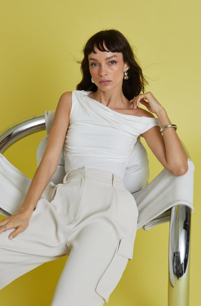 A woman is wearing a sleeveless white top with a gathered, draped design across the front and high-waisted, loose-fitting beige pants with belt loops and a cargo pocket on the side. She accessorizes with pearl earrings, a silver bracelet, and a ring on her finger.