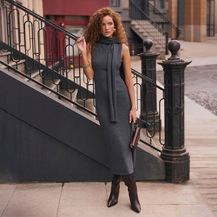 Woman wearing a dark gray sleeveless dress and matching scarf, black boots, holding a handbag.