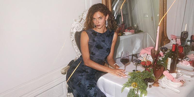 Woman in a sleeveless, dark blue, floral dress with lace accents, sitting at a decorated table.