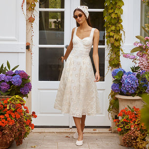 Woman in a white, sleeveless, lace midi dress with floral patterns, paired with white shoes, standing outdoors near potted plants and a white door.