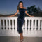 The woman is wearing an off-the-shoulder, dark blue velvet dress that is fitted and reaches below her knees. She is also wearing open-toed high heel shoes with a metallic or shiny finish.