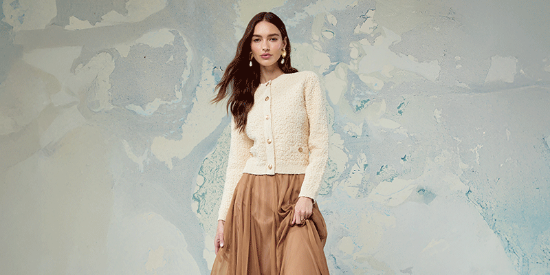 Woman wearing a cream textured button-up cardigan and a long, brown pleated skirt.
