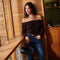 Woman in a black off-the-shoulder top with long sleeves and blue jeans, holding a black handbag.