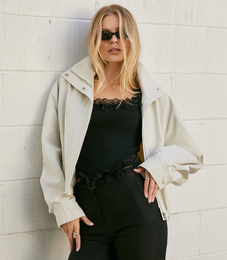 Woman wearing black lace-trimmed top, black pants, and a beige jacket.