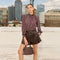 Woman in plaid shirt and black leather shorts holding a brown handbag, urban skyline background.