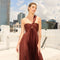 Woman in a one-shoulder, pleated brown dress on a rooftop with city buildings in the background.