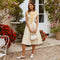Young woman in cream dress with a yellow sweater tied around her shoulders, white shoes, holding a small white bag.