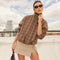 Woman wearing a brown plaid long-sleeve shirt with a beige skirt, sunglasses, and holding a handbag.