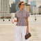 Woman in checkered top and white wide-leg pants, holding a pink coffee cup and a brown handbag.