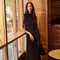 Woman in a sleeveless black dress holding a black handbag, standing indoors by a window.