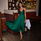 Woman in a flowing emerald green dress with spaghetti straps, black high heels, and a gold purse.
