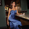 Young woman in a shiny blue satin slip dress with ruffle detail and thin straps, sitting at a bar.