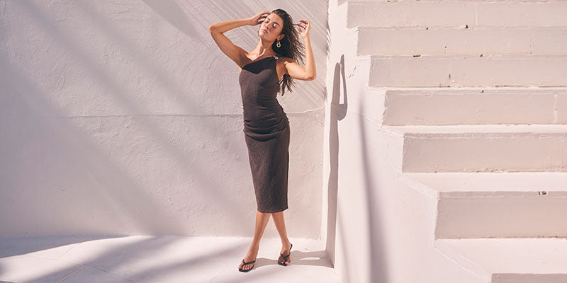 Woman in a fitted, dark brown sleeveless midi dress and black high heels standing by stairs.