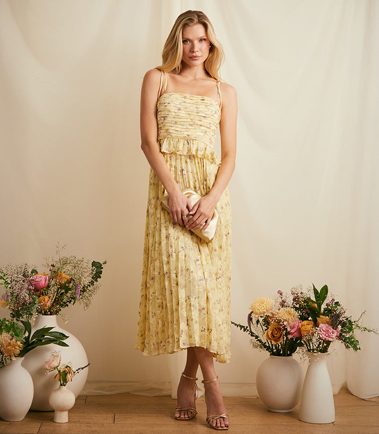 Woman in a yellow, floral, sleeveless dress with spaghetti straps and a pleated skirt.