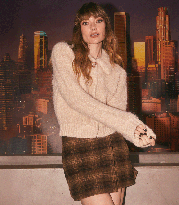 Woman in a beige, fuzzy sweater tucked into a brown plaid skirt with a side slit, standing in front of a cityscape backdrop.