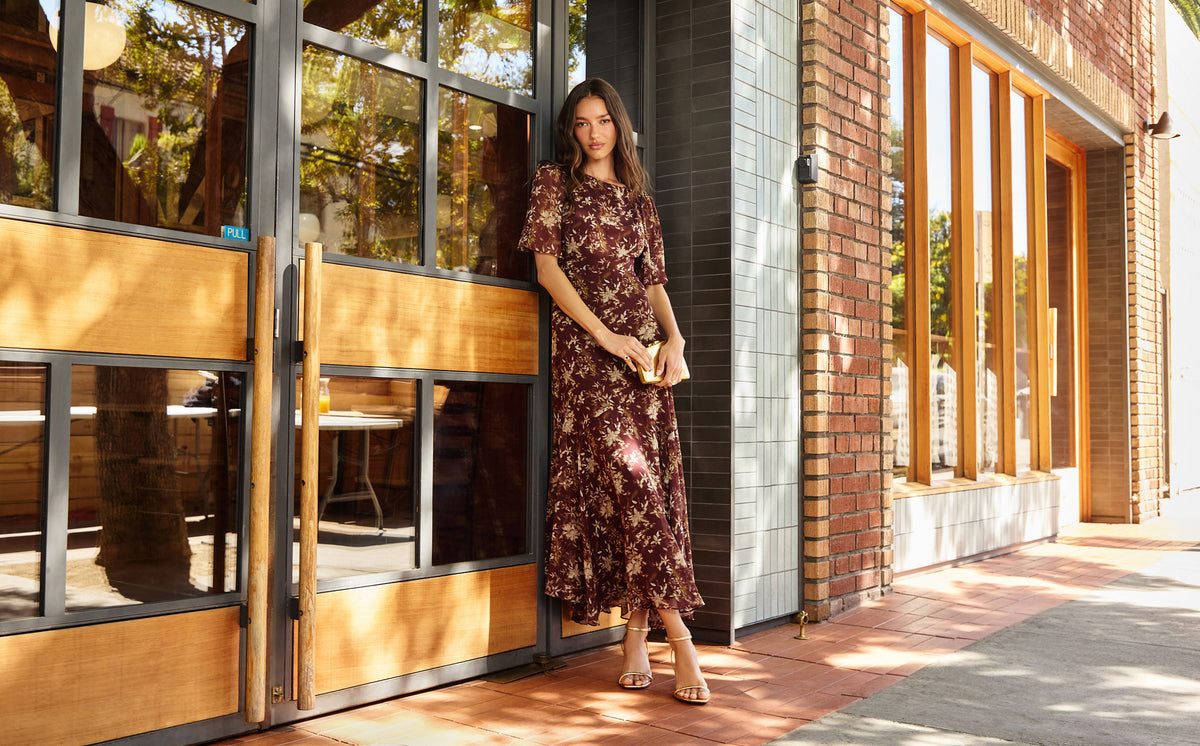 Woman in a long, dark red floral dress standing outside a building with large wooden and glass doors.