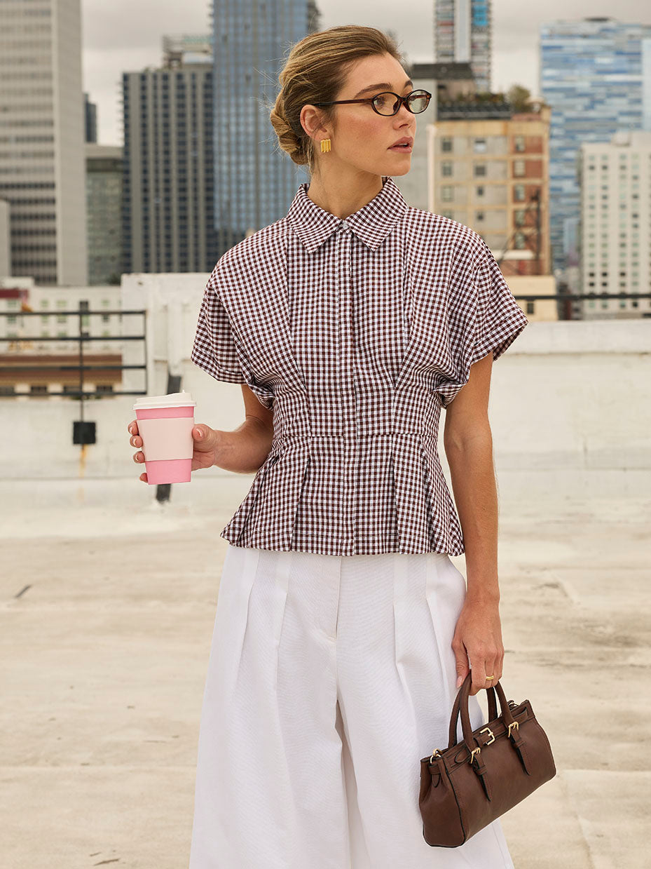 Woman wearing a short-sleeved, checkered blouse with a collar and pleated white wide-leg pants, holding a pink coffee cup and a brown handbag.
