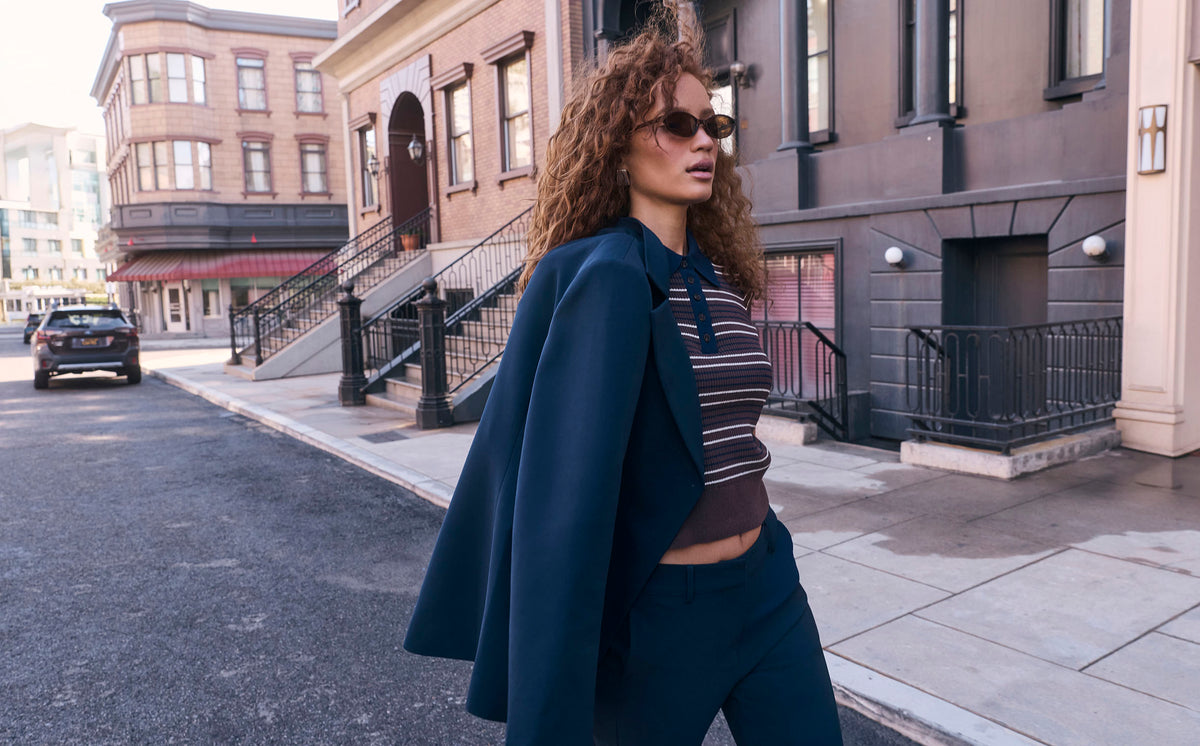 Woman wearing a dark blue blazer over a striped crop top and dark blue pants, with sunglasses.