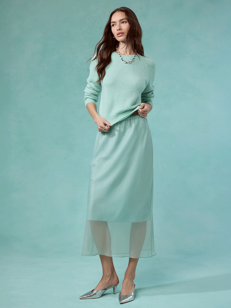 Woman wearing a mint green sweater, satin midi skirt with a sheer overlay, and silver heels.