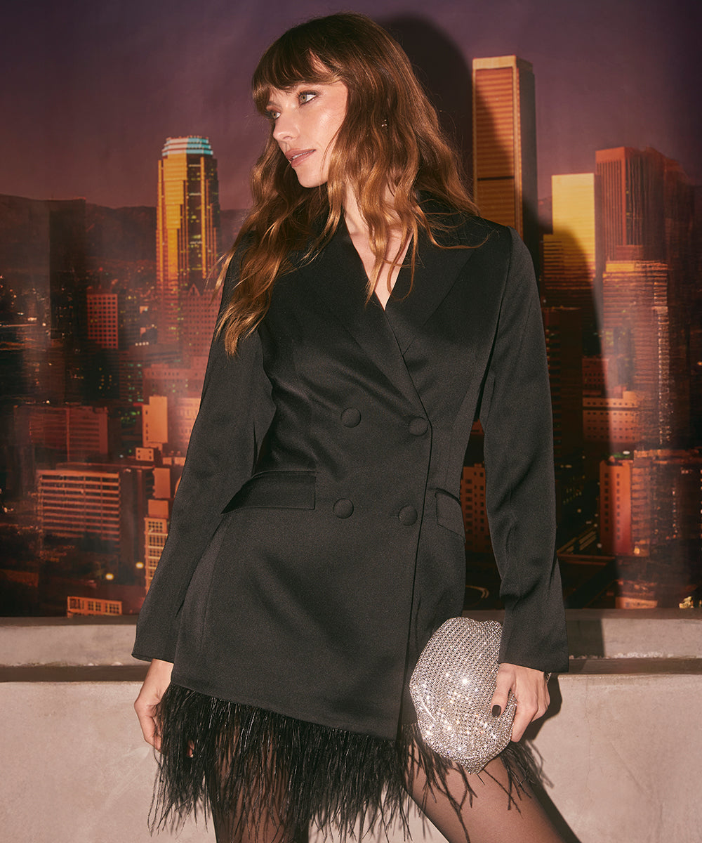 A woman is wearing a tailored black blazer with a double-breasted design, featuring buttons and a structured fit. The blazer is paired with a skirt or dress that has black feathers at the hem, adding a textured detail. She is holding a small, shiny, bejeweled clutch bag.