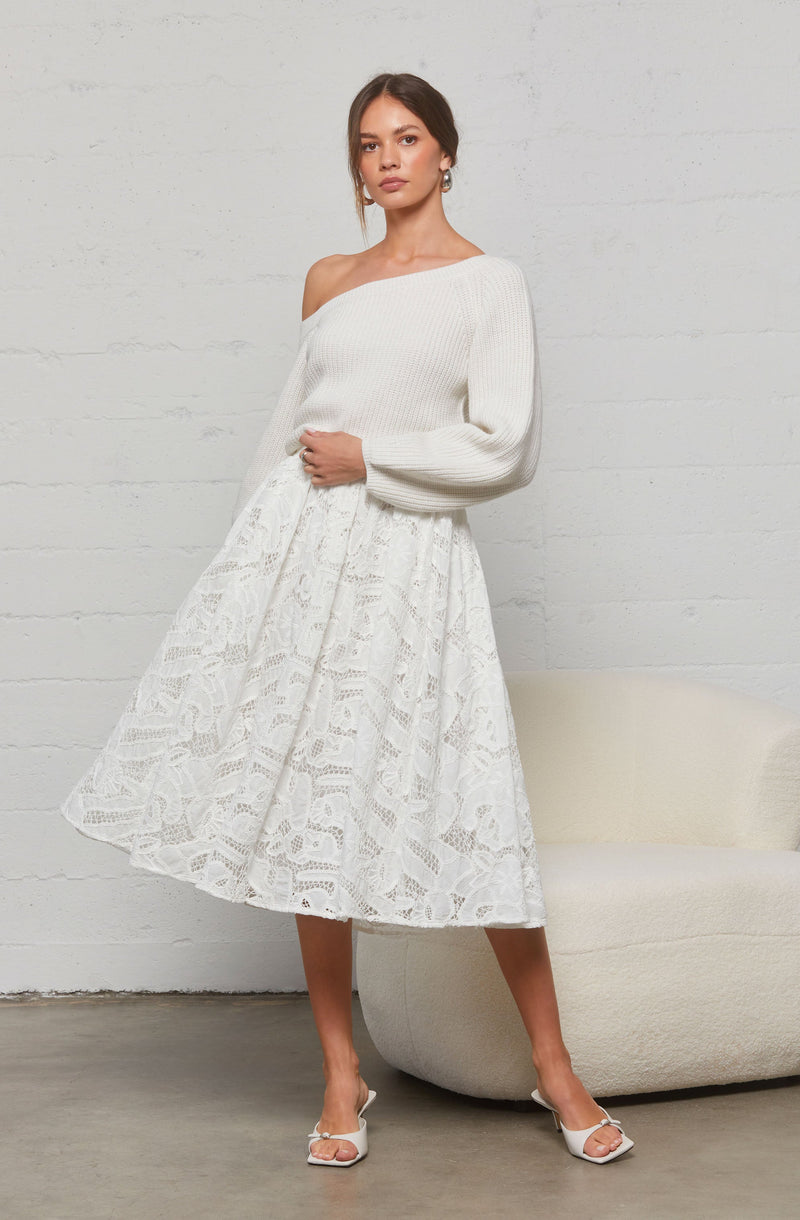 A woman is wearing an off-the-shoulder, long-sleeved, white knit sweater paired with a white, knee-length lace skirt featuring a floral and geometric pattern. She is also wearing white high-heeled shoes with ankle straps.