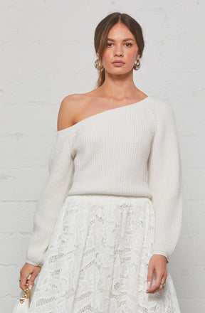 A woman is wearing a white, off-the-shoulder knit sweater with long sleeves. She has a matching white lace skirt that features intricate lace patterns. She accessorizes with large hoop earrings and rings on her fingers.