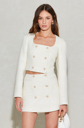 A woman is wearing a white, textured two-piece outfit consisting of a long-sleeved, cropped jacket with a rounded neckline and decorative buttons down the front, and a matching high-waisted mini skirt with similar buttons. She accessorizes with gold hoop earrings and holds a white handbag with a gold chain strap.