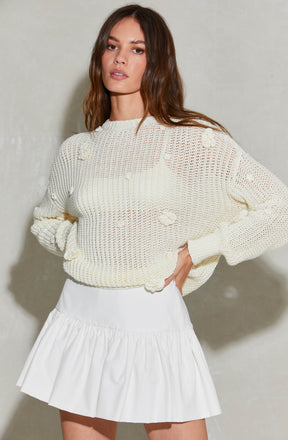 A woman is wearing a loose-knit, long-sleeved cream sweater with a distressed, worn look featuring small holes and frayed edges. She pairs it with a white, high-waisted, ruffled skirt that has a gathered, pleated design at the hem.