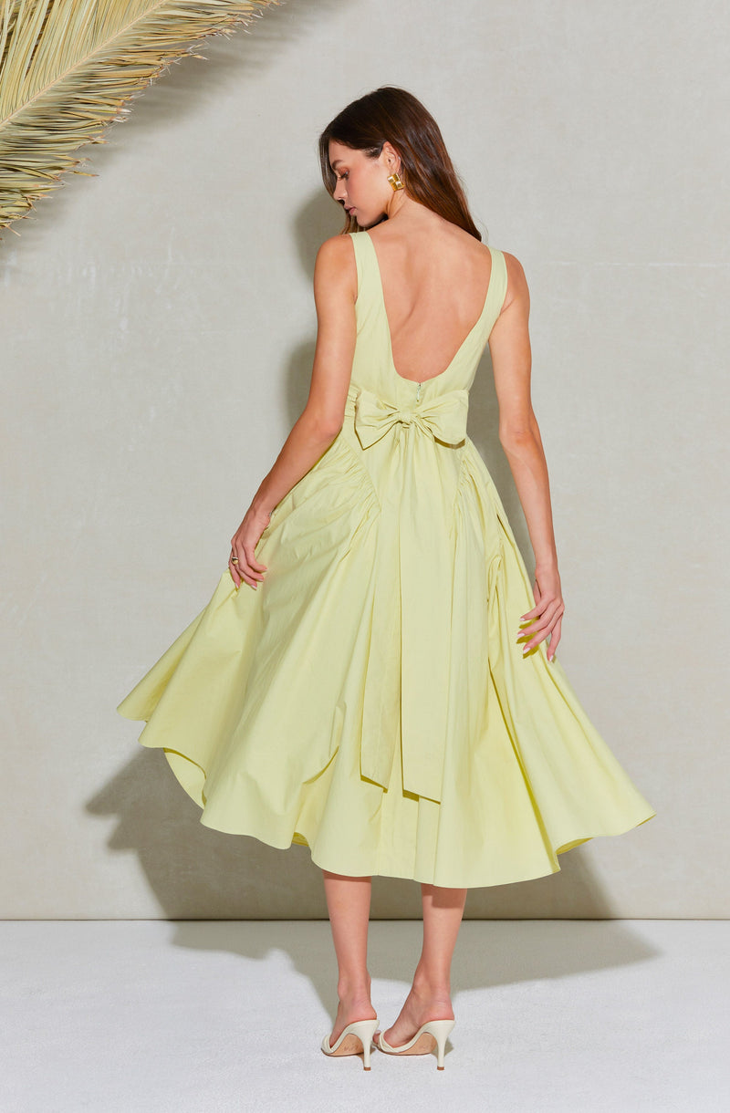 A woman is wearing a sleeveless, light yellow dress with a deep scoop back. The dress has a fitted bodice with wide straps and a large bow at the back. The skirt is voluminous and midi-length, with pleats and drapes that create a flowing silhouette. She is also wearing white high-heeled shoes and gold earrings. The background is plain with a neutral tone, and a large palm leaf is in the top left corner.
