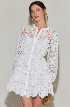 A woman is wearing a white lace dress with floral patterns. The dress features long sleeves, a button-up front, and a waist tie. The lace has a delicate, intricate floral design, and the hemline is scalloped. She is accessorized with gold hoop earrings and holds a small woven bag.