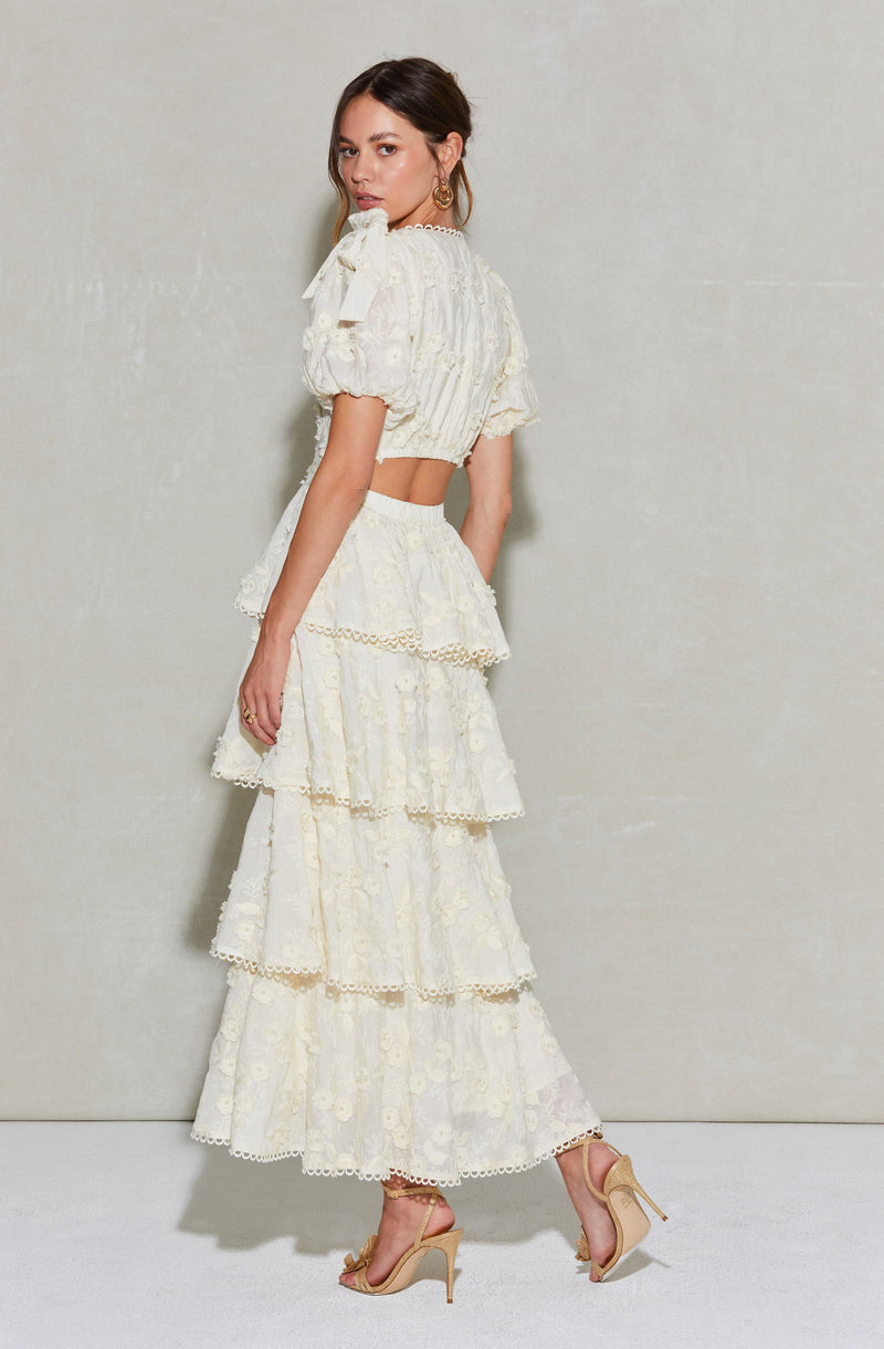 A woman is wearing a two-piece cream-colored outfit with intricate lace and embroidery details. The top has puffed sleeves with bows on the shoulders and a cropped length. The matching skirt is high-waisted, flowing, and features multiple tiered layers with decorative trim at the edges. She is also wearing beige high-heeled sandals with ankle straps.