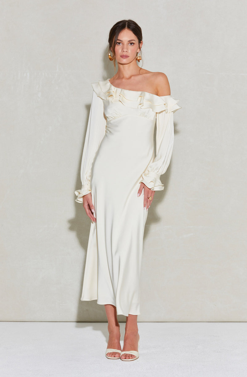 A woman is wearing an off-the-shoulder, cream-colored, full-length dress with ruffle detailing around the neckline and ruffled cuffs on the long sleeves. The dress is fitted and flows down to the floor. She is accessorized with large, gold hoop earrings and is wearing light-colored open-toe heels.