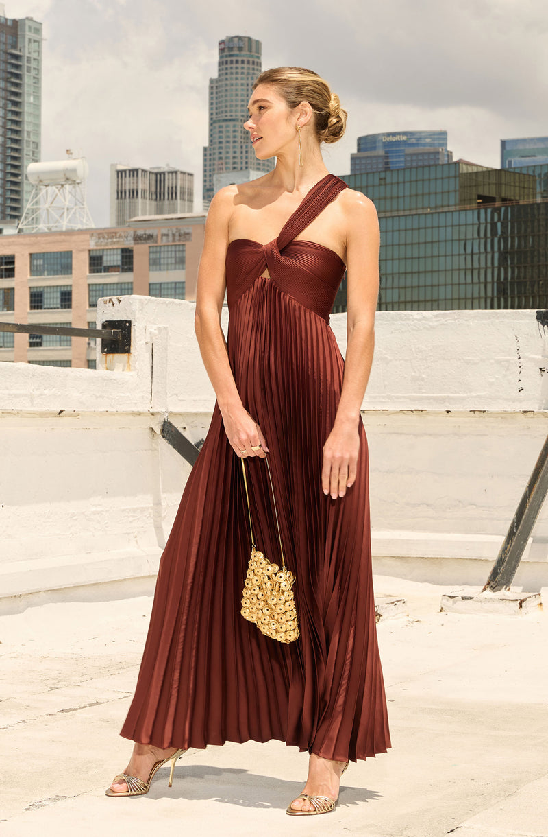 Woman in a one-shoulder, pleated, brick-red dress with gold accessories on a rooftop.