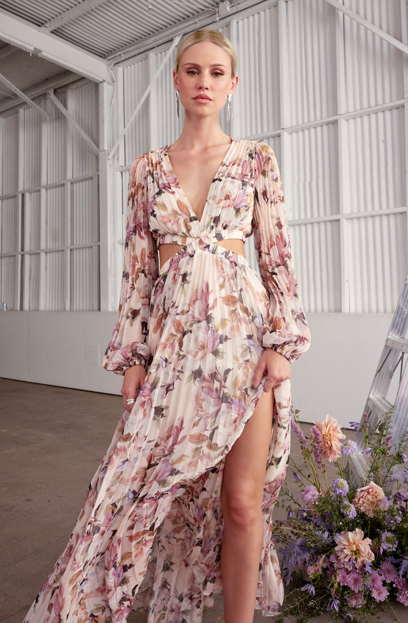 A woman is wearing a long, flowy dress with a floral print in soft pink, purple, and beige tones. The dress features a deep V neckline, long sheer sleeves with elastic cuffs, and has cut-outs at the waist. The skirt of the dress has a high slit revealing her leg. The fabric is lightweight and slightly pleated, creating a delicate and elegant appearance.